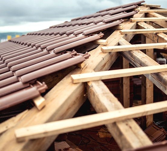 Pose de tuiles brunes sur une charpente en bois neuve ou en rénovation, sous un ciel gris, montrant la structure du toit.