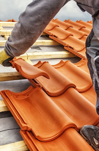 Couvreur ganté en train de poser des tuiles en terre cuite rouge-orange sur une charpente de toit, en cours de construction.