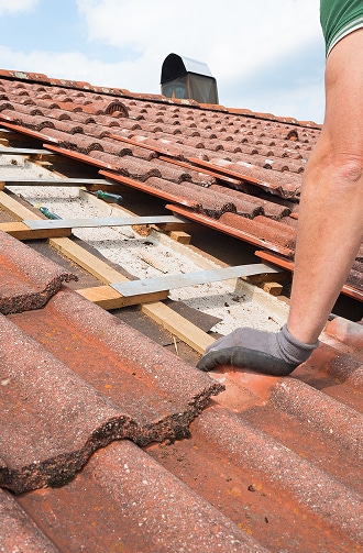 Un couvreur avec un gant gris travaille sur un toit en tuiles rouges, soulevant des tuiles pour réparer la sous-toiture et les lattes.