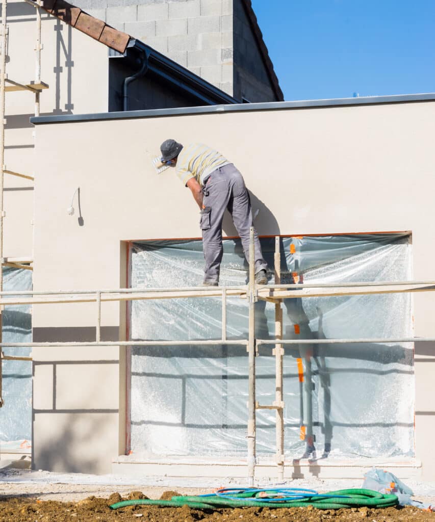 Crépis de façade : l'application de l'enduit par l'ouvrier. Ouvrier sur échafaudage appliquant l'enduit de finition beige sur la façade d'un bâtiment en construction. Fenêtres protégées.