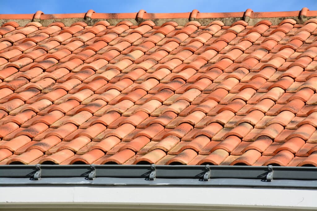 Toit en tuiles terre cuite : Gouttière et couverture Toit ancien en tuiles canal de terre cuite orange-rouge sous ciel bleu. Gouttière métallique grise fixée à l'avant-toit blanc.